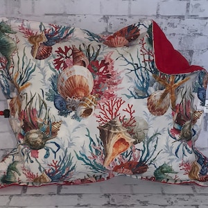 May include: A white cushion with a red border features a colourful seashell and coral design. The cushion is decorated with a variety of seashells, coral, and starfish.