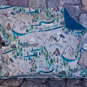 May include: A gray and blue fabric pillow with a map design featuring mountains, rivers, and trees. The text on the pillow reads "Over the Mountains", "Along the River", "Into the Woods", and "Into the Woods".