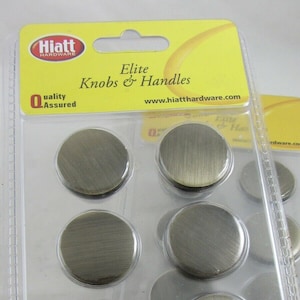 Può includere: Un pacchetto di otto pomelli per armadi in ottone anticato con viti incluse. La confezione è etichettata "Hiatt Hardware Elite Knobs & Handles" e "Quality Assured".