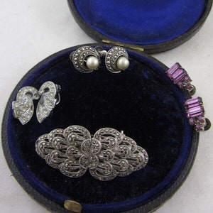 Op de afbeelding: Een blauwe fluwelen juwelendoos met een zilveren broche, twee paar oorbellen en een paar paarse strass oorbellen. De doos heeft een gouden inscriptie met de tekst "J.W. Benson Ludgate Hill London."