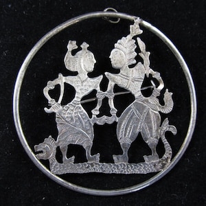 Puede incluir: Un colgante de plata con un diseño circular que presenta dos figuras que se toman de la mano. Las figuras están intrincadamente detalladas y parecen estar bailando. El colgante está rodeado por un marco circular.