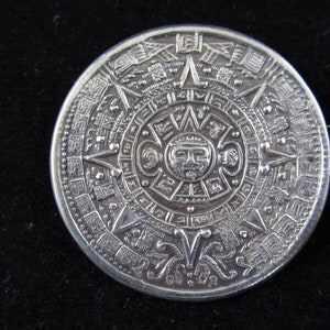 May include: A silver pendant featuring a detailed depiction of the Aztec calendar, also known as the Sun Stone. The circular pendant is adorned with intricate carvings and symbols.