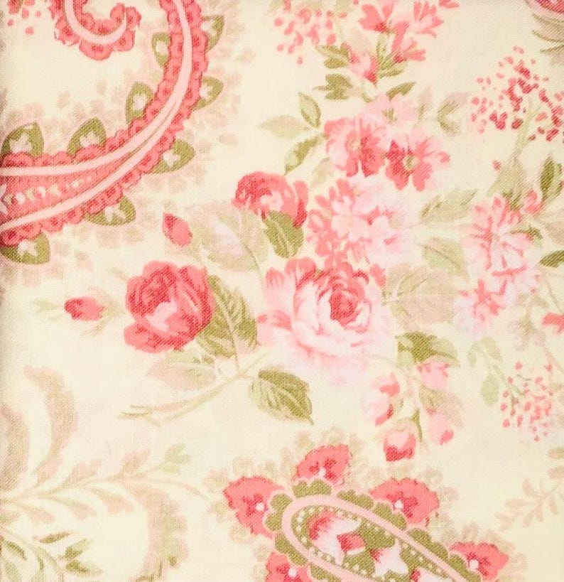May include: A floral fabric featuring a cream background with a pattern of pink roses, smaller pink flowers, and green leaves. The design includes paisley-like shapes in pink and green, creating a vintage, romantic aesthetic.
