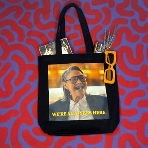 May include: A black canvas tote bag featuring a printed image of a person wearing glasses and a white shirt under a dark blazer. The text WE'RE ALL DYKES HERE is printed in yellow below the image. The bag contains a small book, a gold pen, and dried lavender.