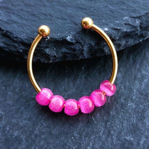 Könnte beinhalten: Ein goldfarbener, offener Ohrring mit sechs rosa Perlen. Die Enden des Ohrrings haben kleine, runde goldfarbene Akzente. Der Ohrring ist vor einem dunklen, strukturierten Hintergrund platziert.