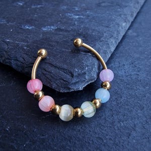 Op de afbeelding: Een goudkleurige manchetarmband met een rij kleurrijke, doorschijnende kralen in roze, geel en blauw. De armband heeft een eenvoudig, open ontwerp met kleine gouden bollen aan elk uiteinde. De kralen worden gescheiden door kleine gouden kralen.