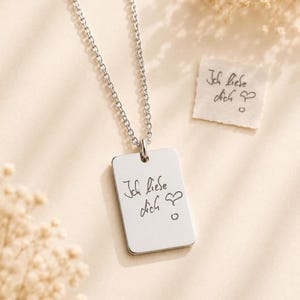 Puede incluir: Un collar con colgante rectangular plateado con la frase manuscrita « Ich liebe dich » grabada. El collar está colgado de una cadena plateada. También se ve un pequeño trozo de papel con la misma frase.