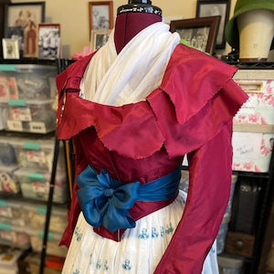 以下が含まれることがあります： フリルネックと長袖の、バーガンディ色の歴史的衣装のボディス。ウエストには、青緑色のサッシュとリボンがアクセントになっています。白いスカートには、繰り返しの青い模様があります。衣装はマネキンに展示されています。