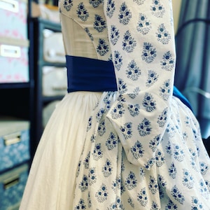 Puede incluir: Vestido blanco con mangas y detalles de corpiño con estampado floral azul. El vestido presenta una faja ancha azul oscuro en la cintura y una falda amplia. Las mangas tienen una ligera forma de campana. El vestido parece un disfraz histórico o un diseño de inspiración vintage.