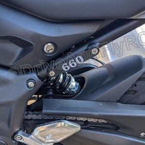 Puede incluir: Primer plano de una motocicleta negra con una cadena plateada y un guardabarros negro. La motocicleta tiene un amortiguador plateado con un resorte negro y un tornillo plateado con el n&uacute;mero "660" impreso en &eacute;l.