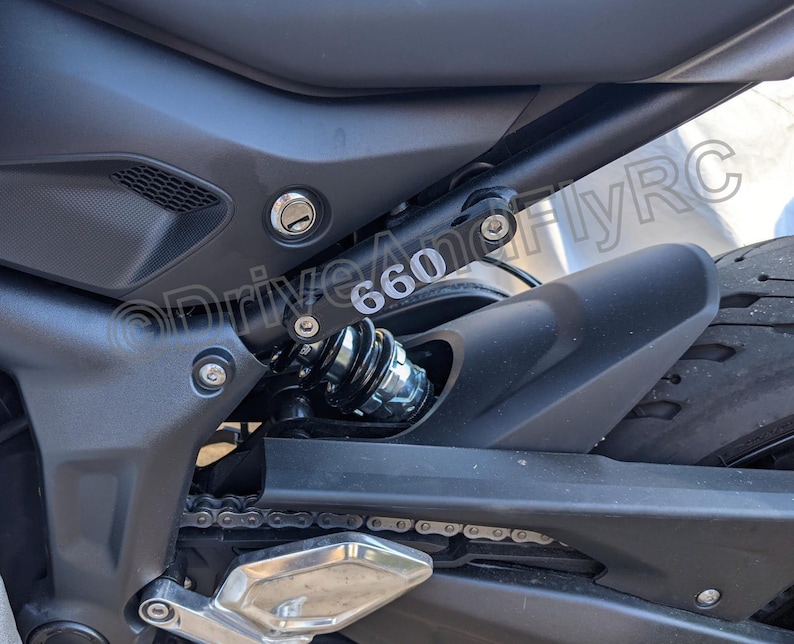 Puede incluir: Primer plano de una motocicleta negra con una cadena plateada y un guardabarros negro. La motocicleta tiene un amortiguador plateado con un resorte negro y un tornillo plateado con el n&uacute;mero "660" impreso en &eacute;l.