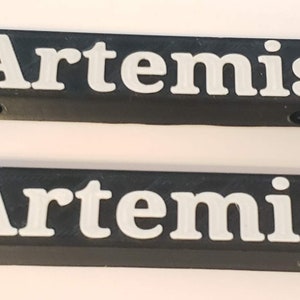 Puede incluir: Dos placas de nombre de pl&aacute;stico negro con la palabra "Artemis" impresa en letras blancas.