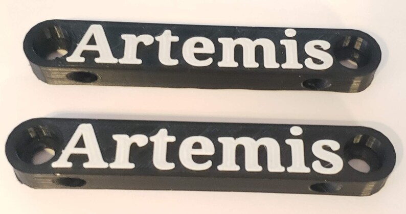 Puede incluir: Dos placas de nombre de pl&aacute;stico negro con la palabra "Artemis" impresa en letras blancas.