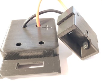May include: A black plastic 3D printed box with two holes on the top and a slot on the bottom. A black plastic connector with a red and black wire is attached to the box.