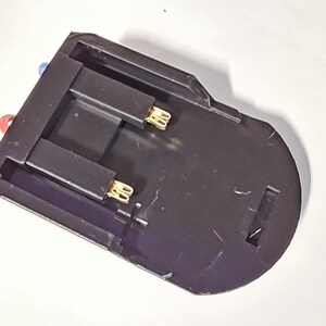 K&ouml;nnte beinhalten: Schwarzer Batteriehalter aus Kunststoff mit zwei goldfarbenen Kontakten. Der Halter hat ein rotes und ein blaues Kabel.