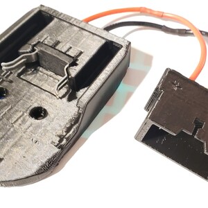 May include: A black 3D printed battery holder with a red and black wire attached to a black connector. The battery holder has a slot for a battery and two metal clips.