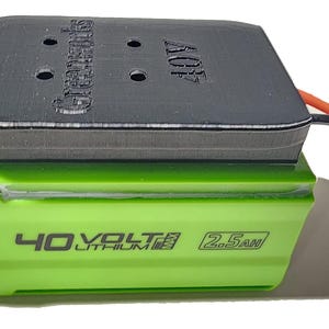 May include: A green and black 40V lithium battery with a black adapter. The battery has the text "40 VOLTS LITHIUM 2.5AH" printed on it. The adapter has the text "Greenworks" and "40V" printed on it. An orange and black wire is connected to the adapter.
