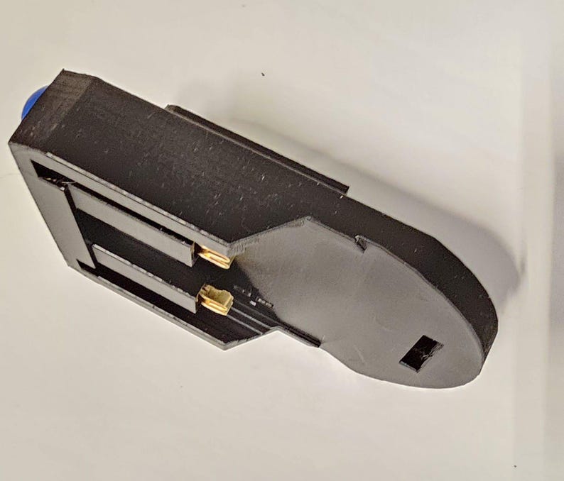 K&ouml;nnte beinhalten: Ein schwarzer Batteriehalter aus Kunststoff mit zwei vergoldeten Kontakten. Der Halter hat einen blauen Knopf oben.