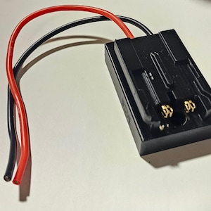 May include: A black electronic component with gold-colored connectors and two wires, one red and one black. The wires extend from the component, which has a rectangular shape with recessed areas.