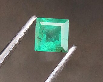 Puede incluir: Una esmeralda verde vibrante, de corte cuadrado, es sostenida por unas pinzas plateadas sobre un fondo borroso. Las facetas de la esmeralda reflejan la luz, mostrando su claridad y color. Esta gema es un hermoso ejemplo de esmeraldas naturales.
