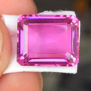 Puede incluir: Una gema rosa rectangular y facetada, con un aspecto pulido. La gema se presenta sobre una superficie blanca, resaltando su claridad y color. El corte de la piedra crea múltiples facetas, reflejando la luz y realzando su brillo.