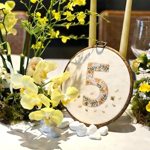 May include: A wooden embroidery hoop table number with the number 5 embroidered in a floral pattern. The number is surrounded by yellow flowers, white stones, and two tall candles.