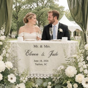 Może przedstawiać: Biały ślubny bieżnik na stół z napisem Mr. & Mrs. Elowen & Jude, June 18, 2026, Taylors, SC. Bieżnik posiada koronkową lamówkę i jest wyeksponowany na stole z dwoma kieliszkami do szampana, w otoczeniu białych kompozycji kwiatowych.