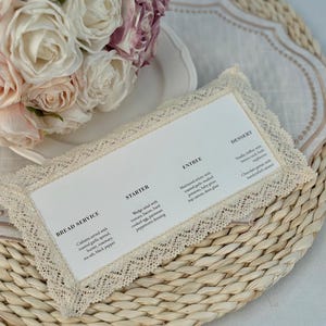 Puede incluir: Una tarjeta de menú con un borde de encaje se encuentra sobre un mantel individual tejido. El menú enumera las opciones de servicio de pan, entrante, plato principal y postre. Un ramo de rosas blancas y rosas está en el fondo.