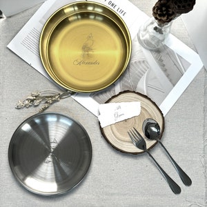 May include: Two metal plates, one gold and one silver, with engraved designs. The gold plate has a bird and the name "Alexander". A fork and spoon are next to a wooden coaster with a handwritten note. Decorative elements complete the scene.