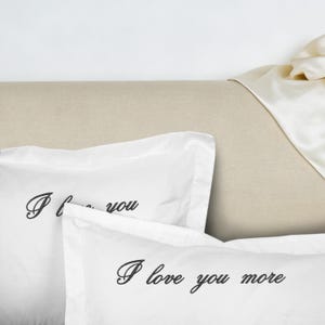 May include: White pillowcases with black script that reads "I love you more" and "I love you". The pillowcases are on a beige surface with a white satin sheet in the background. The pillowcases are folded.