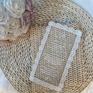 May include: A clear acrylic menu card with a white lace border, featuring menu items in elegant script. The card is placed on a woven placemat, with a bouquet of white and purple roses in the background. The menu includes appetizers, entrees, and desserts.