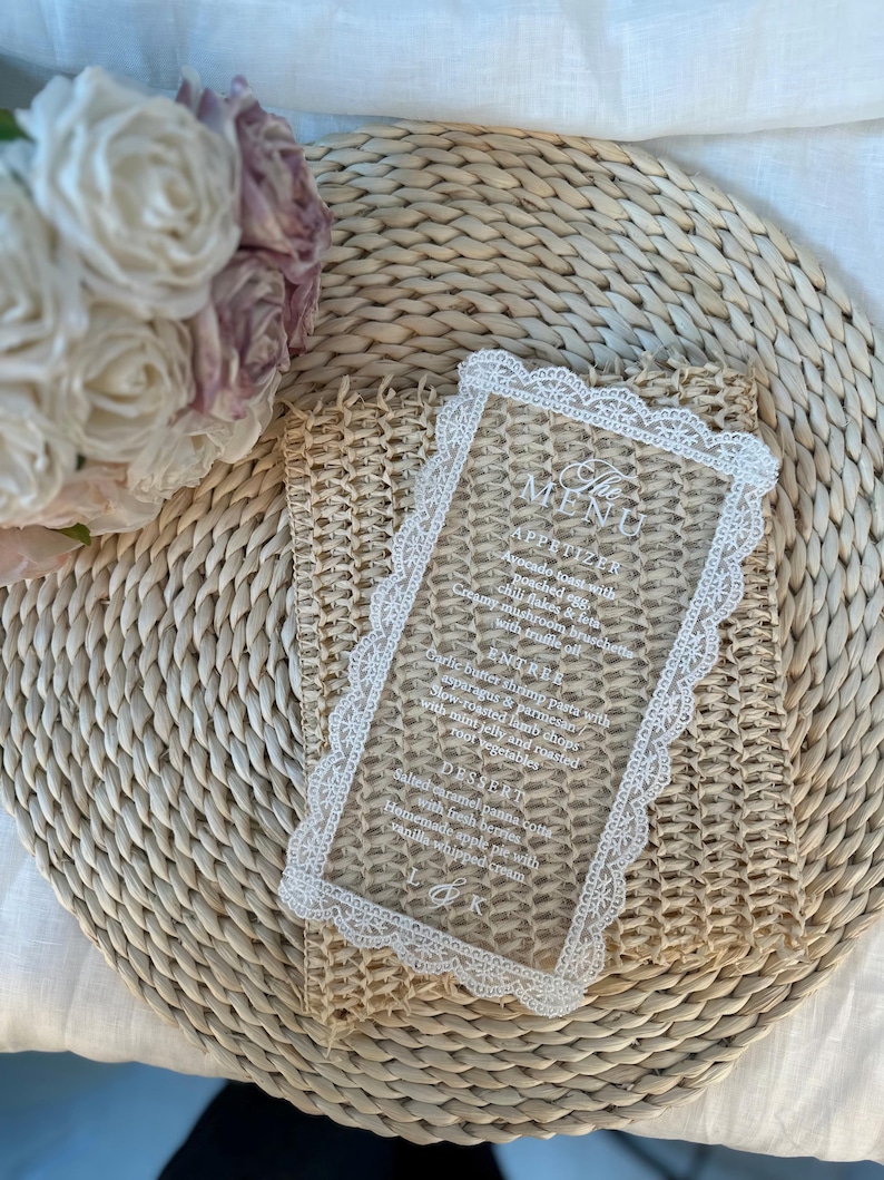 May include: A clear acrylic menu card with a white lace border, featuring menu items in elegant script. The card is placed on a woven placemat, with a bouquet of white and purple roses in the background. The menu includes appetizers, entrees, and desserts.