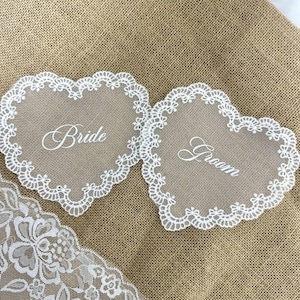 May include: Two heart-shaped wedding decorations with white lace trim. One says "Bride" and the other says "Groom" in elegant script. The decorations are on a burlap background, with a piece of white lace in the corner.