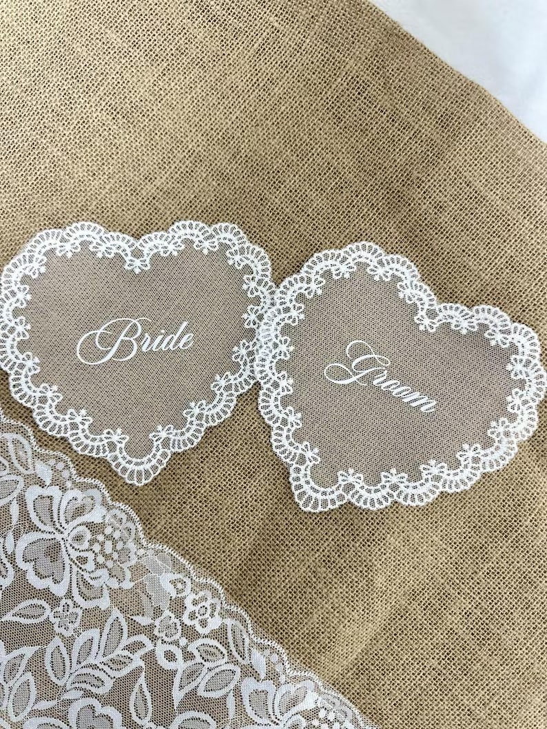 May include: Two heart-shaped wedding decorations with white lace trim. One says "Bride" and the other says "Groom" in elegant script. The decorations are on a burlap background, with a piece of white lace in the corner.