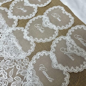 May include: A collection of heart-shaped place cards with white lace edging and personalized names in elegant script. The cards are arranged on a burlap surface, with a white lace floral design visible in the background.