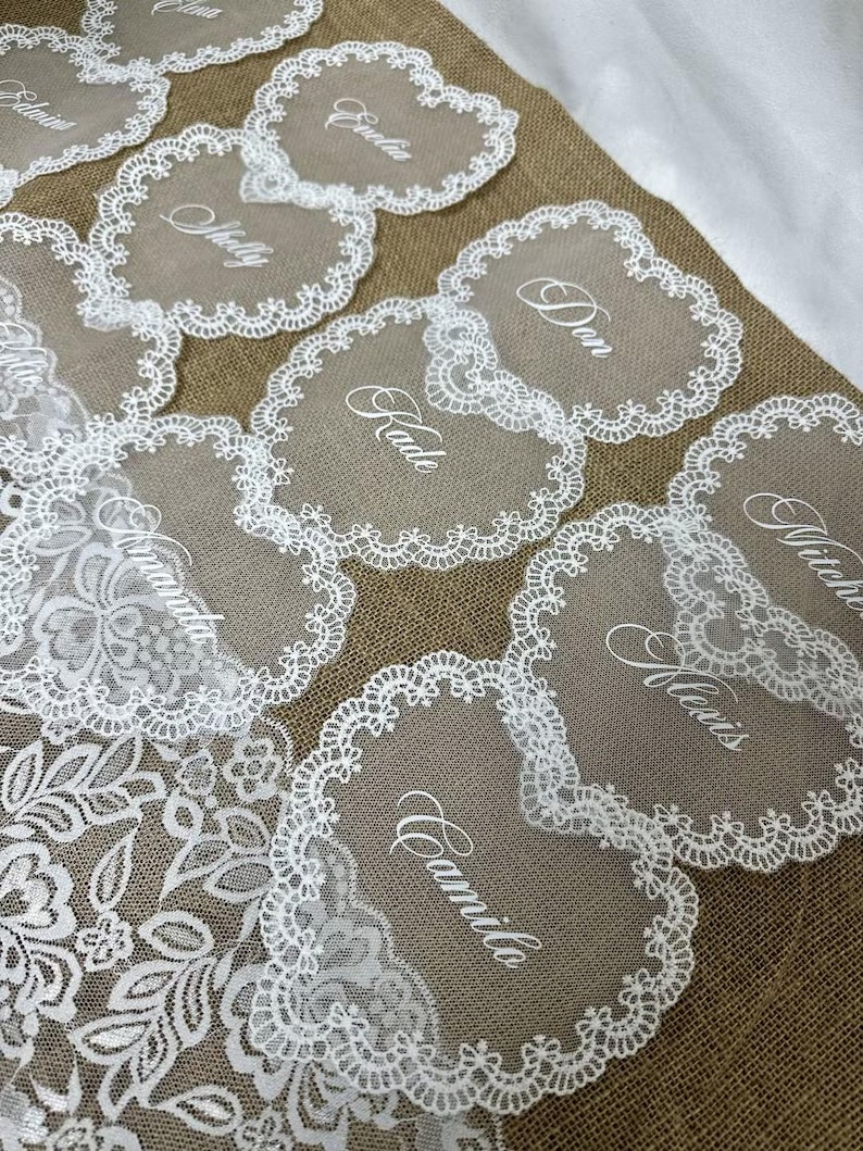 May include: A collection of heart-shaped place cards with white lace edging and personalized names in elegant script. The cards are arranged on a burlap surface, with a white lace floral design visible in the background.
