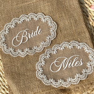Peut inclure: Deux marque-places en acrylique transparent avec bordure en dentelle blanche. L'une des cartes indique "Bride" et l'autre "Miles" en écriture élégante. Les cartes reposent sur une surface en toile de jute, pour mariage.