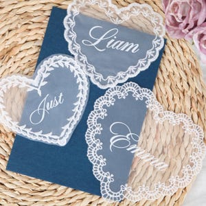 Può includere: Segnaposto a forma di cuore in pizzo con i nomi Liam, Just ed Emma, su un biglietto blu. Il pizzo è bianco con un bordo smerlato. Lo sfondo è un tappetino di paglia intrecciata.