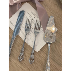 Könnte beinhalten: Ein Set silberner Hochzeitsbestecke, bestehend aus einem Messer, zwei Gabeln und einem Tortenheber, auf einer Holzoberfläche angeordnet. Messer und Tortenheber sind mit Namen und Daten graviert. Eine Hochzeitseinladung und ein rosa Band sind ebenfalls vorhanden.