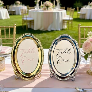 Könnte beinhalten: Zwei ovale Tischschilder, eines goldfarben und eines silberfarben, mit der Aufschrift "Table One" auf cremefarbenem Hintergrund. Sie stehen auf einem rosa Läufer, mit einer verschwommenen Hochzeitsfeier im Freien im Hintergrund.