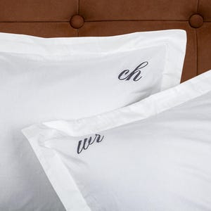 May include: Two white pillowcases with embroidered initials, "ch" and "wr", set against a brown, button-tufted headboard. The pillowcases have a crisp, clean look, suggesting a focus on bedding and home decor.