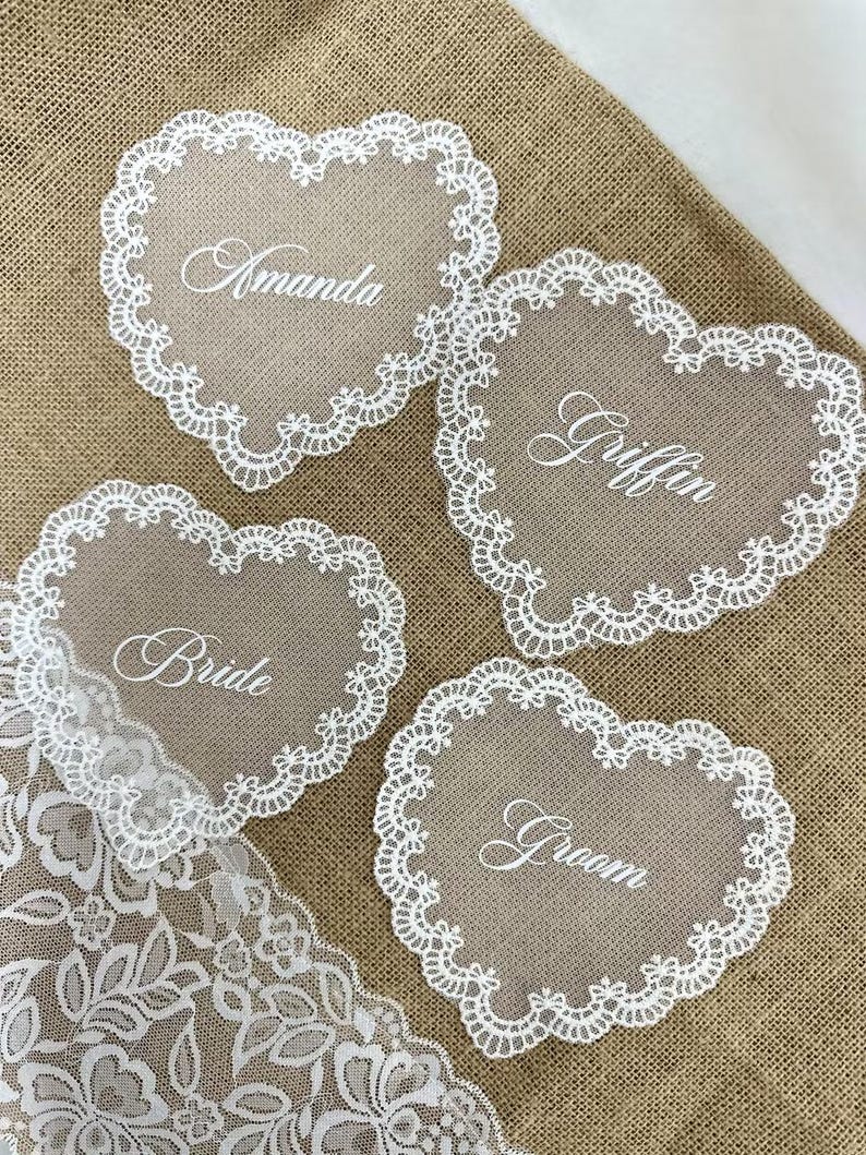 May include: Four heart-shaped place cards with white lace trim and names in cursive: Amanda, Griffin, Bride, and Groom. A white lace trim is visible in the lower left corner. The cards are on a burlap surface.