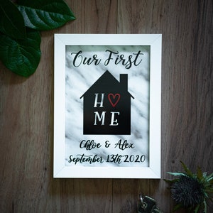 May include: White framed picture with a marble background. The text "Our First Home" is written in black cursive font. A black silhouette of a house with a red heart in the middle is shown. The text "Chloe & Alex September 13th 2020" is written in black cursive font.