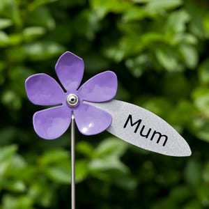 May include: A purple metal flower garden stake with the word "Mum" on a silver leaf-shaped tag. The flower has five petals and a silver centre. The stake is set against a green leafy background.