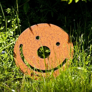 May include: A circular, rusty metal sculpture of a smiling face, featuring two eye holes and a curved mouth. It's placed in a garden amongst green grass and foliage, adding a playful element to the outdoor setting.