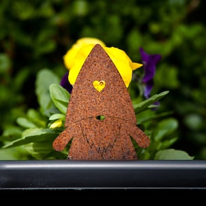 May include: A rusty metal gnome garden decoration with a heart-shaped eye and a beard. The gnome is standing in front of a pot of yellow and purple flowers.
