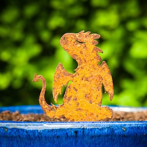 May include: A rusty metal dragon silhouette, with wings and a tail, is placed in a blue ceramic pot filled with brown soil. The dragon is a garden decoration.