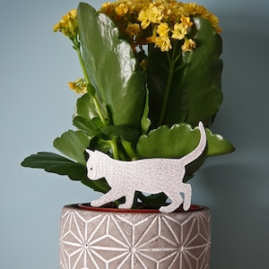 May include: A white ceramic planter with a geometric pattern, featuring a white silhouette of a cat walking across the top. The planter is filled with a green succulent plant with yellow flowers.