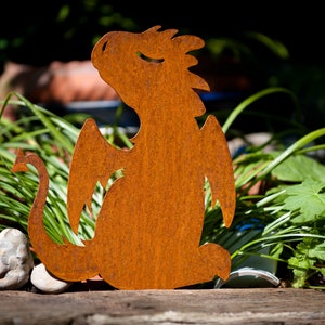 May include: A weathered metal dragon sculpture, featuring a detailed silhouette with wings and a tail. The dragon has a rusty orange finish, set against green plants. This decorative item is suitable for garden or home decoration.