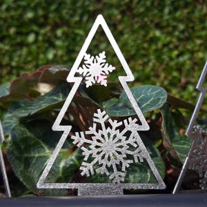 May include: Three silver metal Christmas tree decorations featuring snowflake designs. The decorations are standing upright in a pot of green plants.